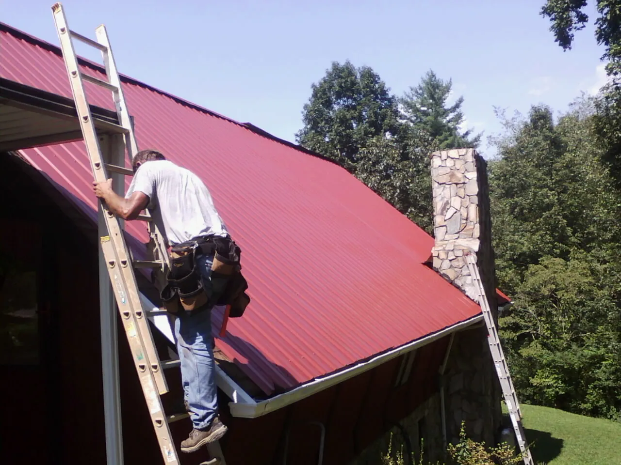 Roofer on a ladder beside a red metal roof and stone chimney — Soffit & Fascia Repair by Roofing Brothers in Finleyville